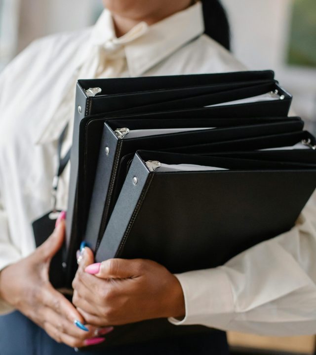Professional woman in formal attire carrying multiple black folders indoors.